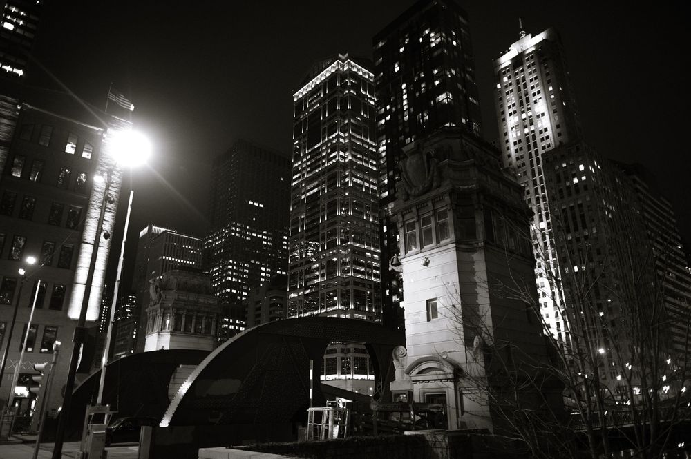 Black and white night cityscape with illuminated skyscrapers and a bright street lamp in an urban downtown setting.