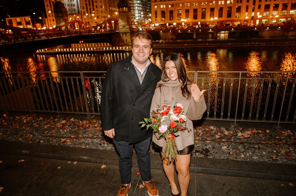 Couple celebrating engagement at night by a river, woman holding red and white flowers, showing ring.