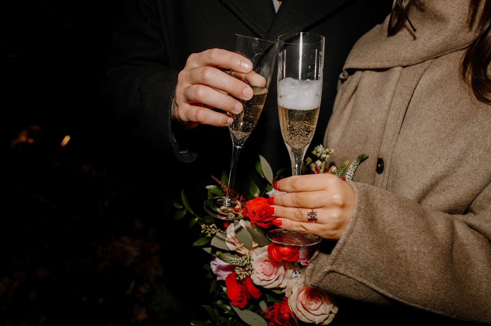 Couple clinking champagne flutes at wedding celebration, bride holding red and pink floral bouquet with engagement ring visible.