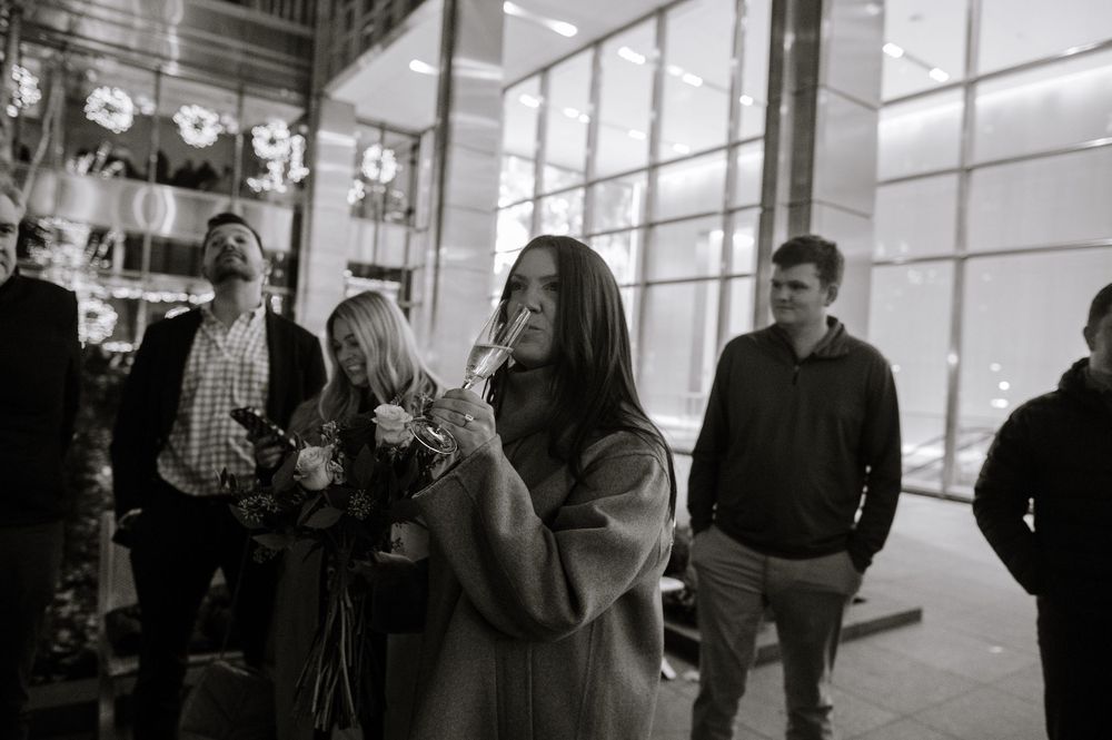 Black and white photo of a woman holding flowers and a drink at an evening event with guests in background.