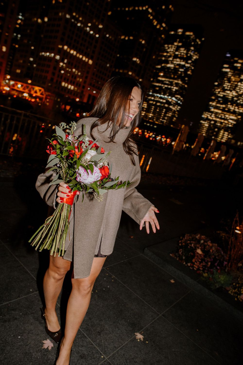 Woman in gray coat holding red flower bouquet smiling at night with city lights in background.