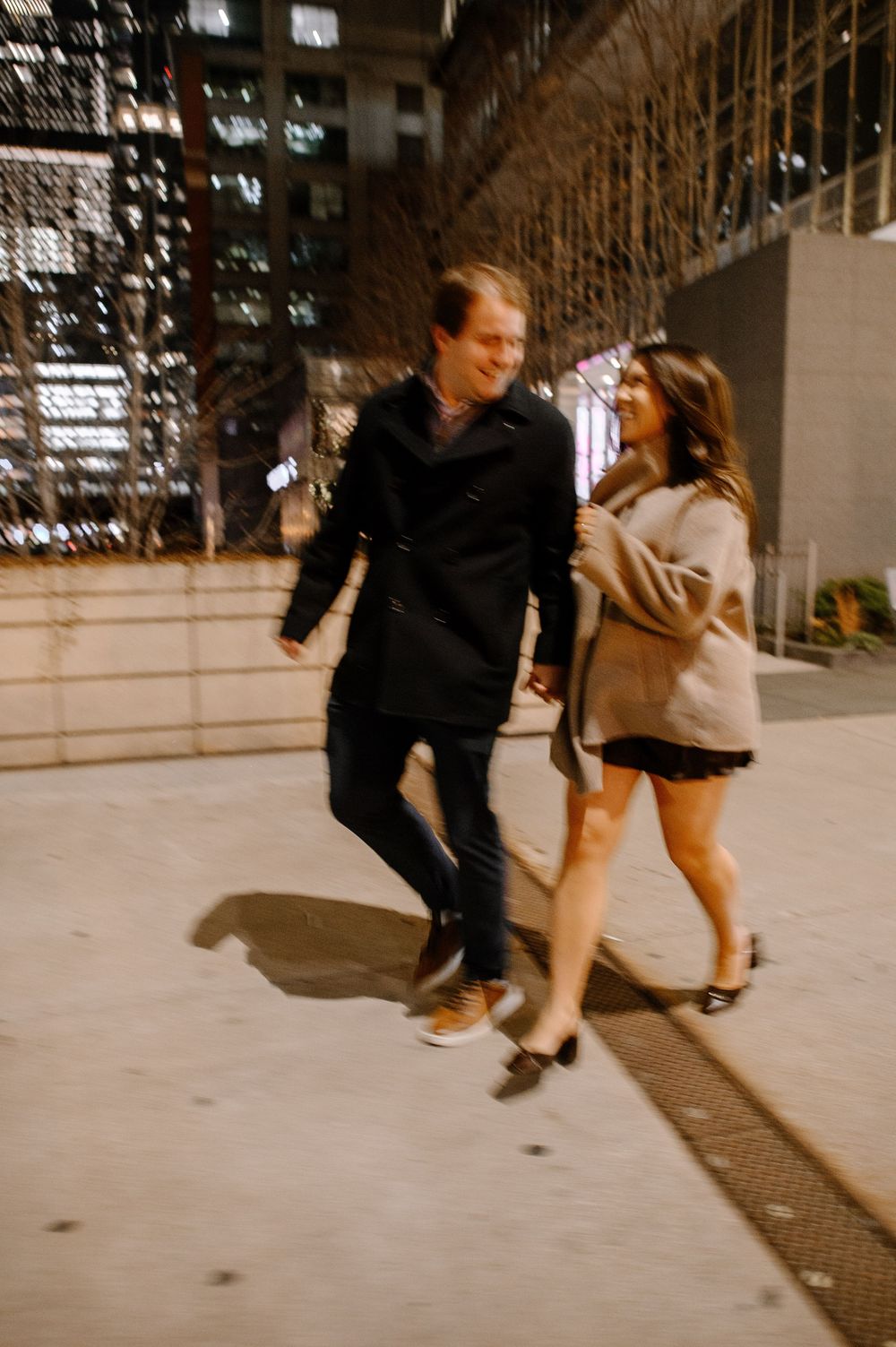 A smiling couple walks hand-in-hand at night in an urban setting, the man in a black coat and woman in a beige sweater.