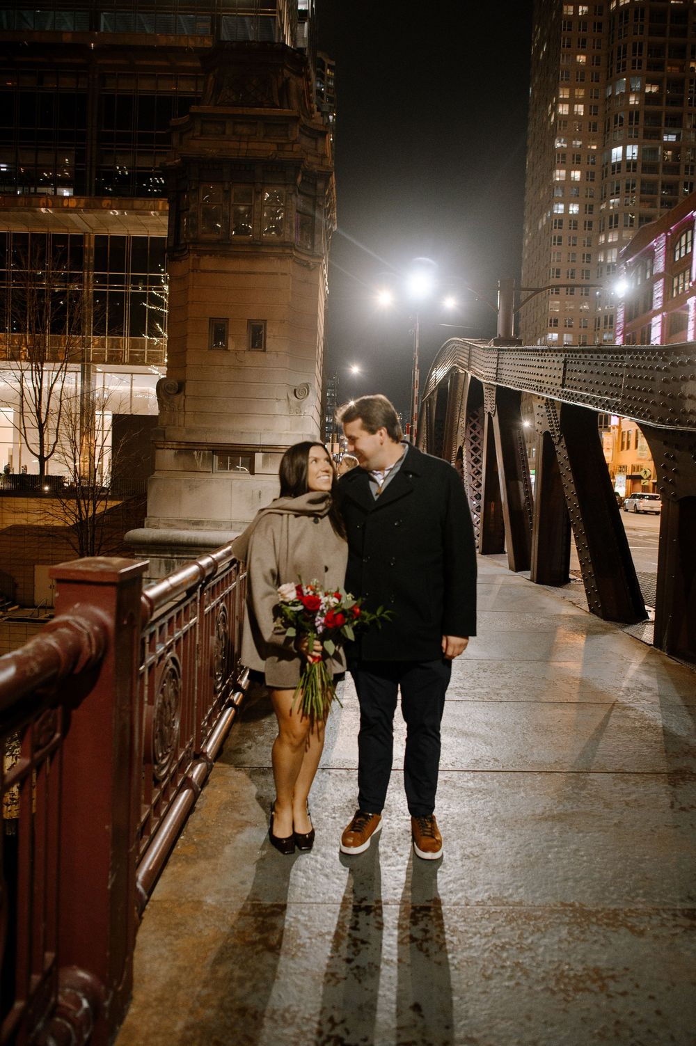 Couple sharing a romantic moment on a Chicago bridge at night, holding red roses with city lights glowing behind them.