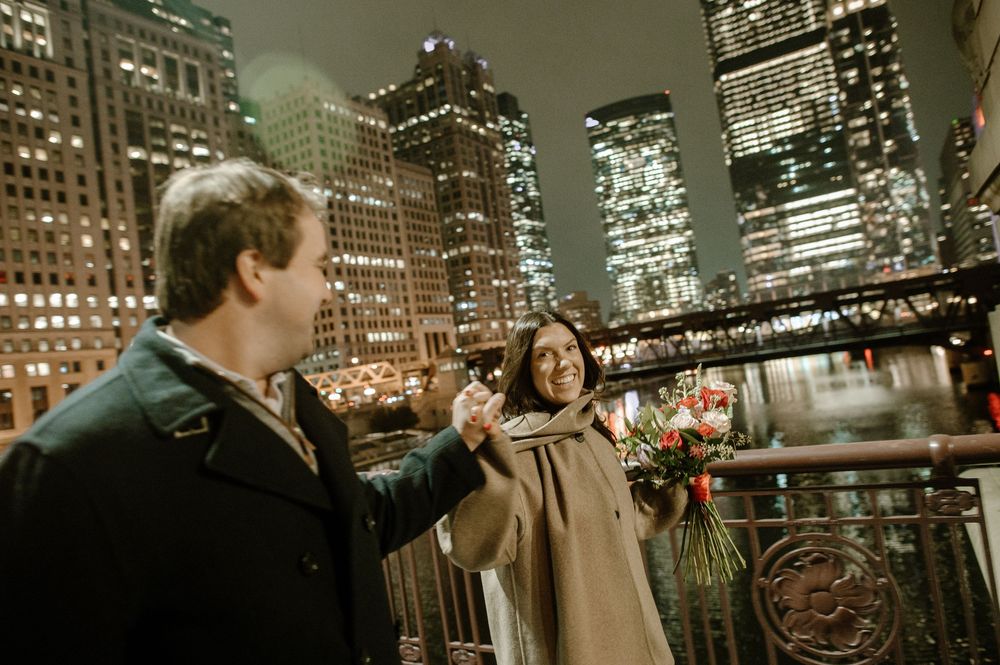 Couple sharing a romantic moment with flowers on a city bridge at night with illuminated skyscrapers.