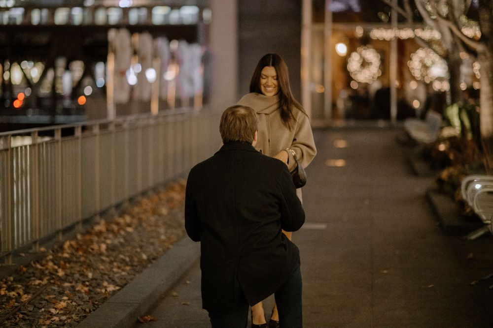 Man kneeling to propose to woman on a city walkway at night with festive lights in background.