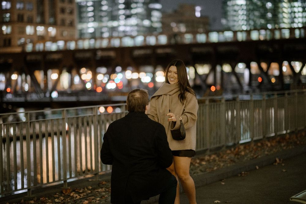 Man proposing to woman on a city bridge at night with glowing urban lights in the background.