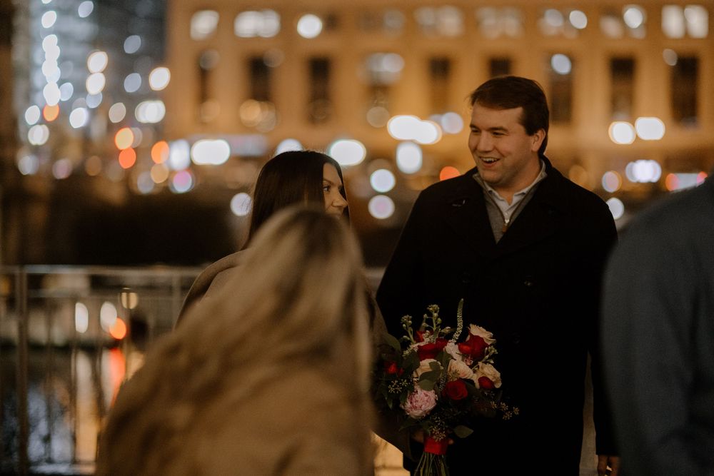 Man holding red flower bouquet smiling at woman outdoors at night with bokeh city lights in background.