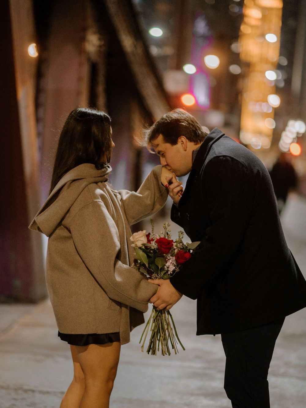 Man kisses woman's hand while holding red roses on a romantic city street at night with bokeh lights.