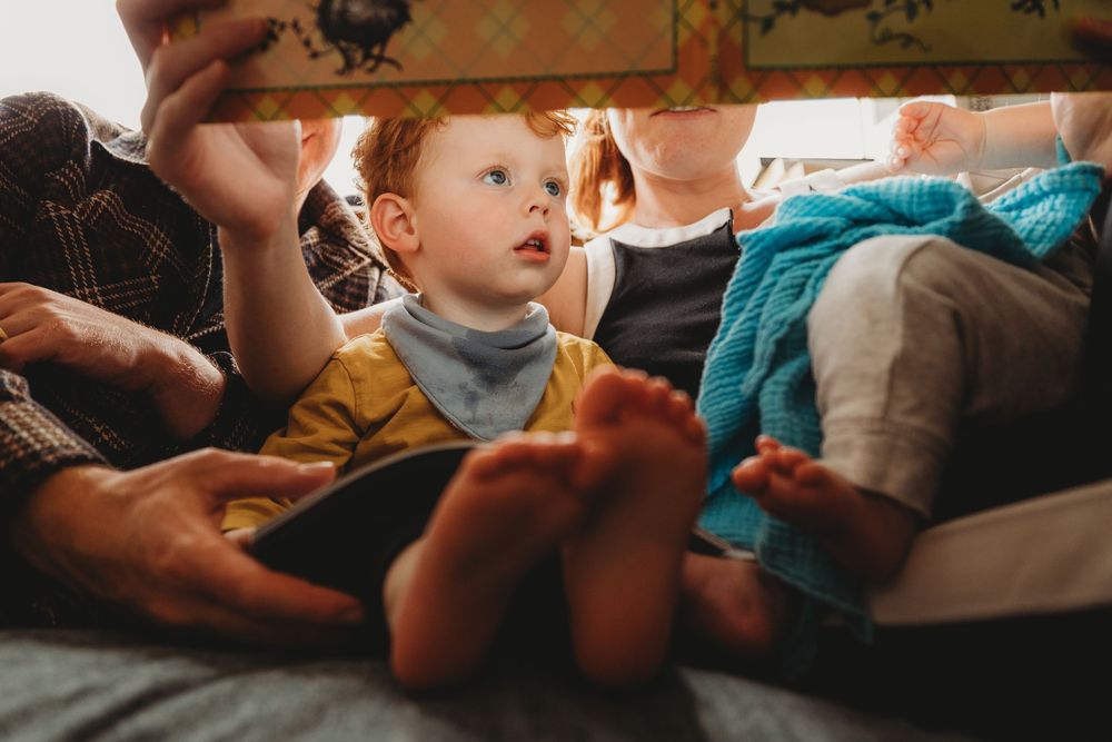 Curious red-haired toddler sitting between adults on a bed, wearing a yellow outfit and bib, looking up with wonder.