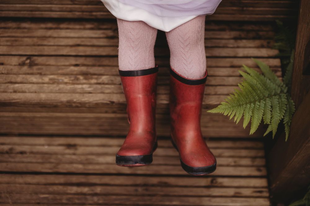 Red rain boots and pink dress shown against rustic wooden planks with green fern leaves in background.