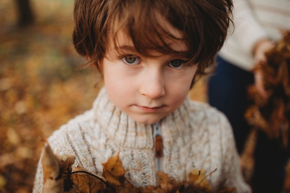 A child with rosy cheeks and dark hair poses outdoors among autumn leaves wearing a cozy knit sweater.