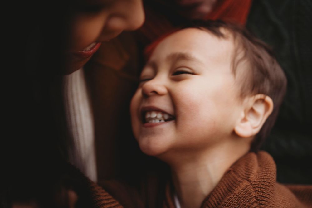 A person wearing a brown knit sweater smiles joyfully against a dark background.