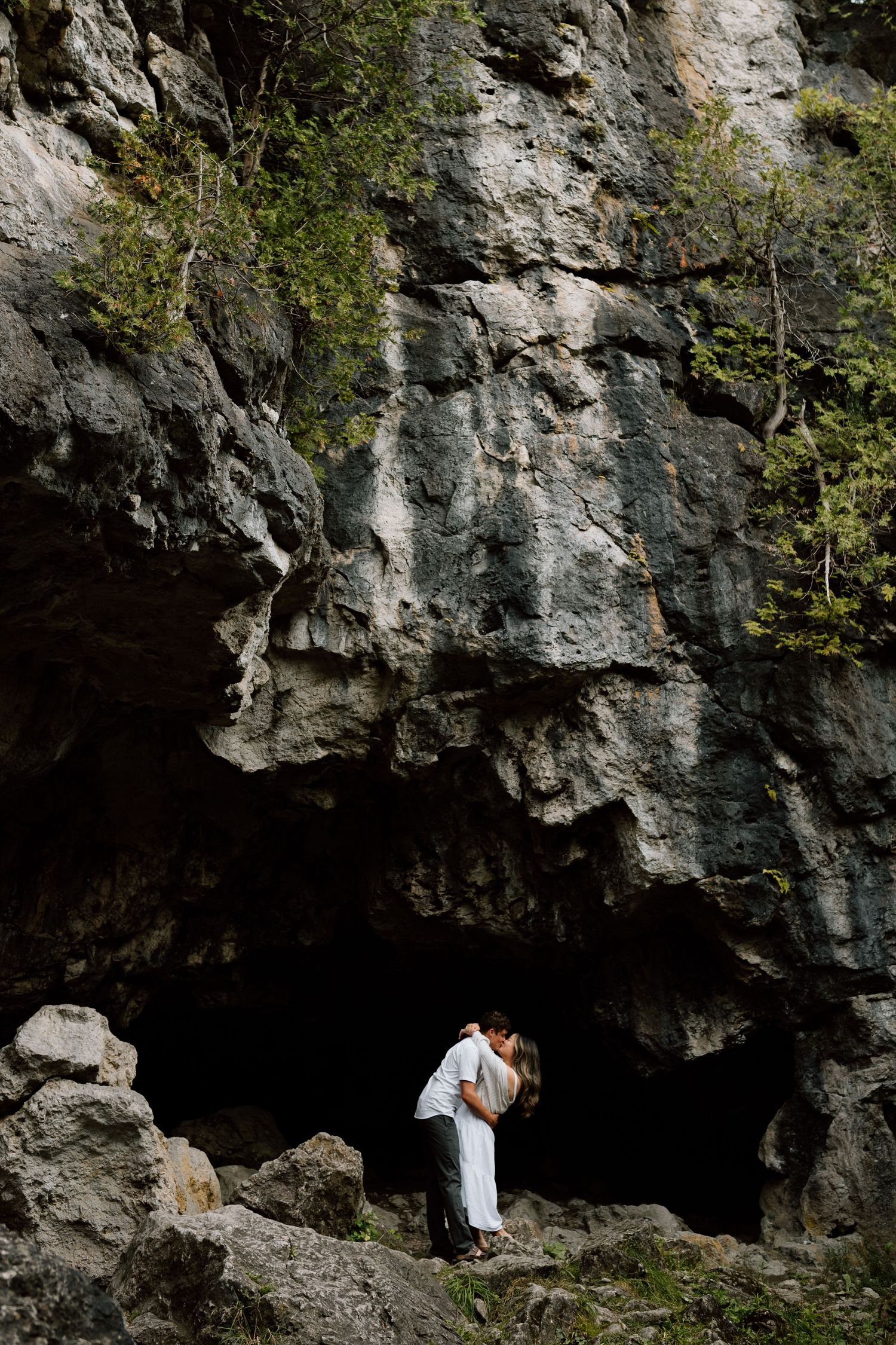 Couple embracing and kissing at the entrance of a dramatic rocky cave surrounded by lush green trees.