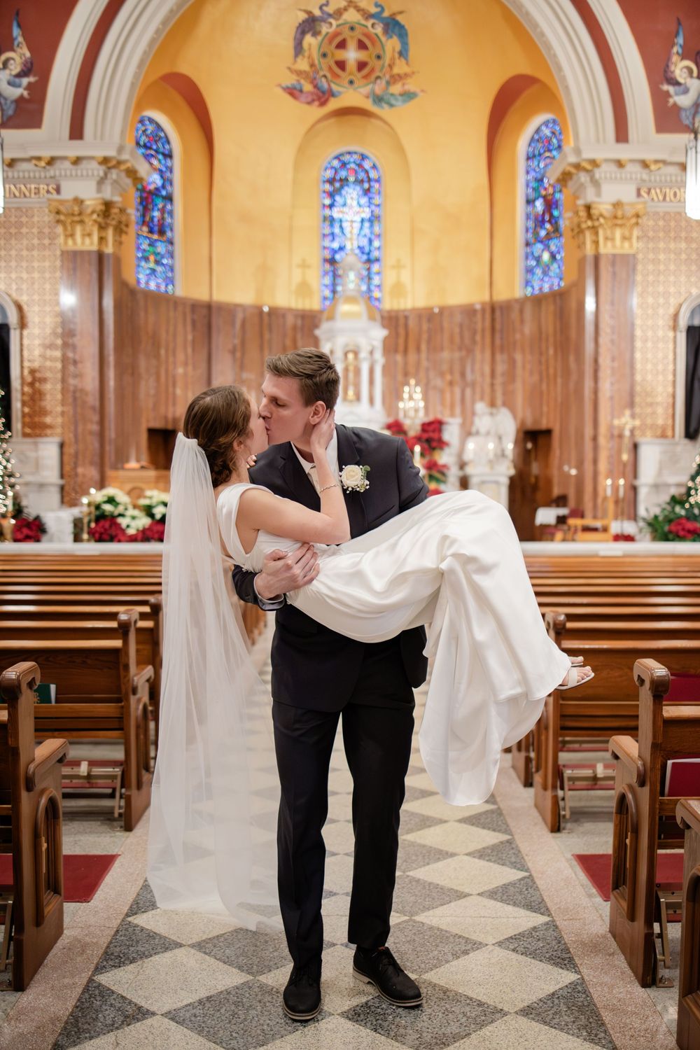 Groom carries bride in his arms while kissing in a beautiful church aisle after wedding ceremony.