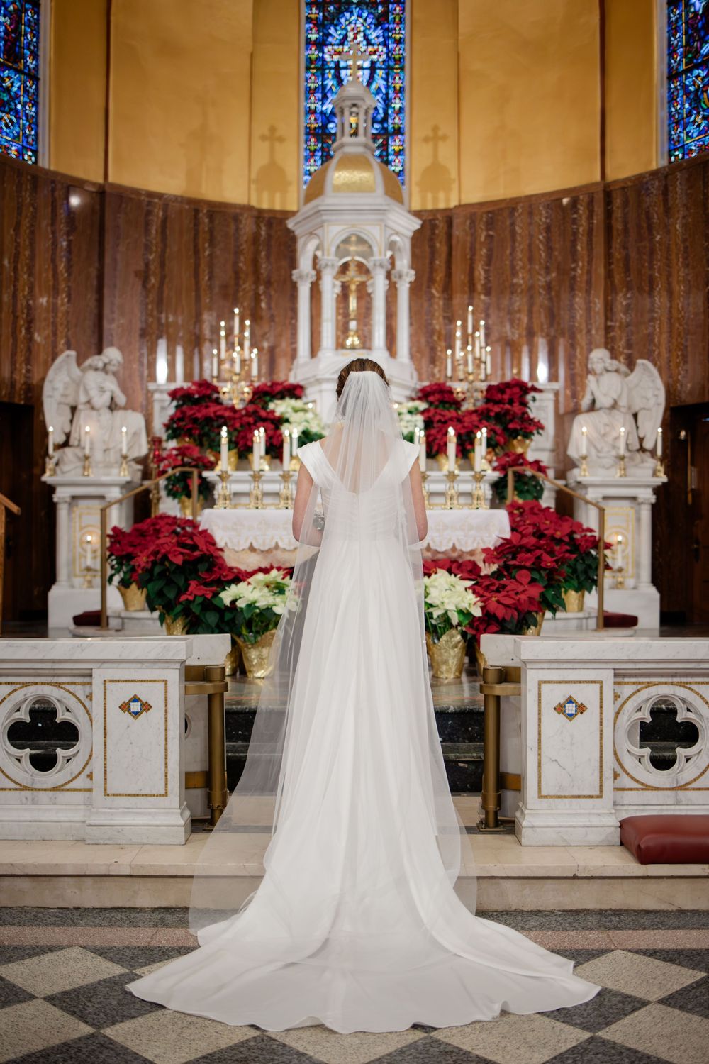 Bride in white gown and veil stands before ornate church altar adorned with red poinsettias and candles.