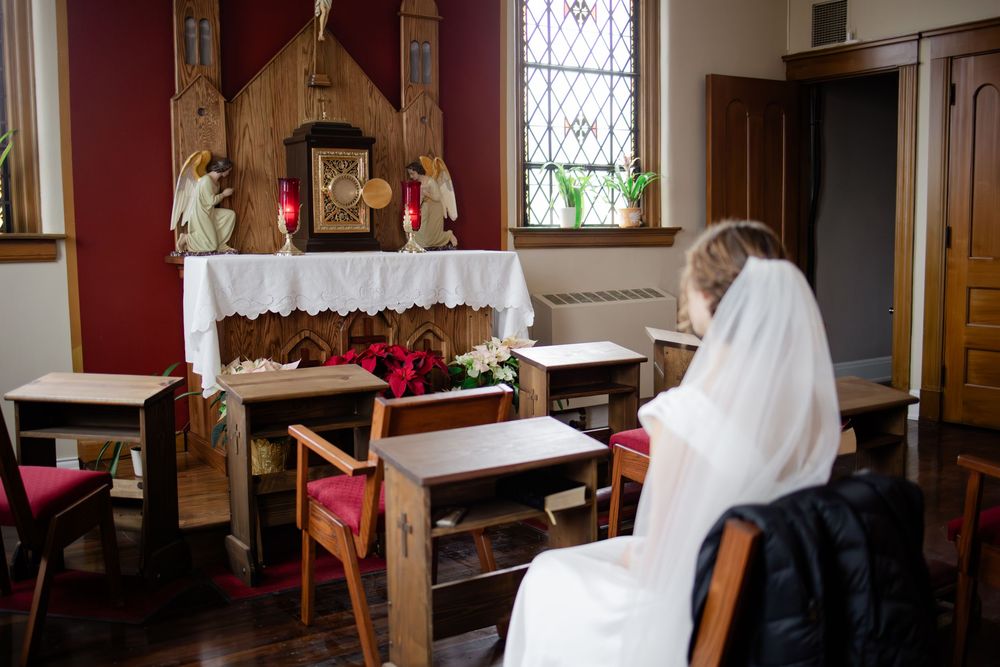 A nun in white habit prays quietly before a wooden altar with candles and religious icons in a chapel.