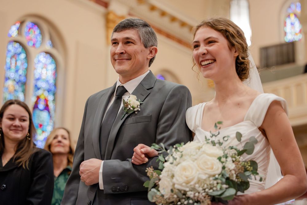 Smiling bride holding white bouquet walks down church aisle with groom in gray suit, stained glass windows behind them.