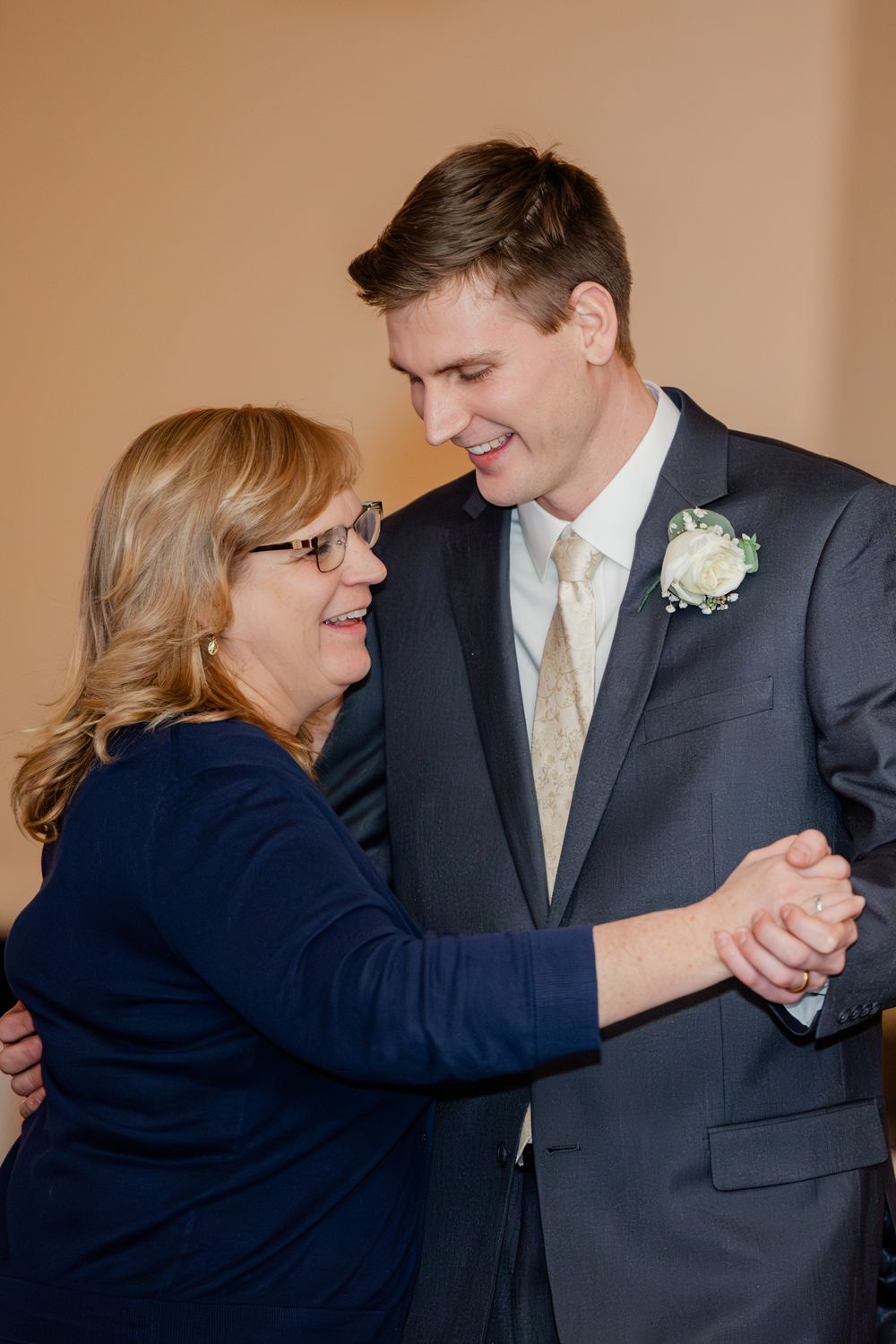 Groom in gray suit dances with smiling mother in navy dress during wedding reception.