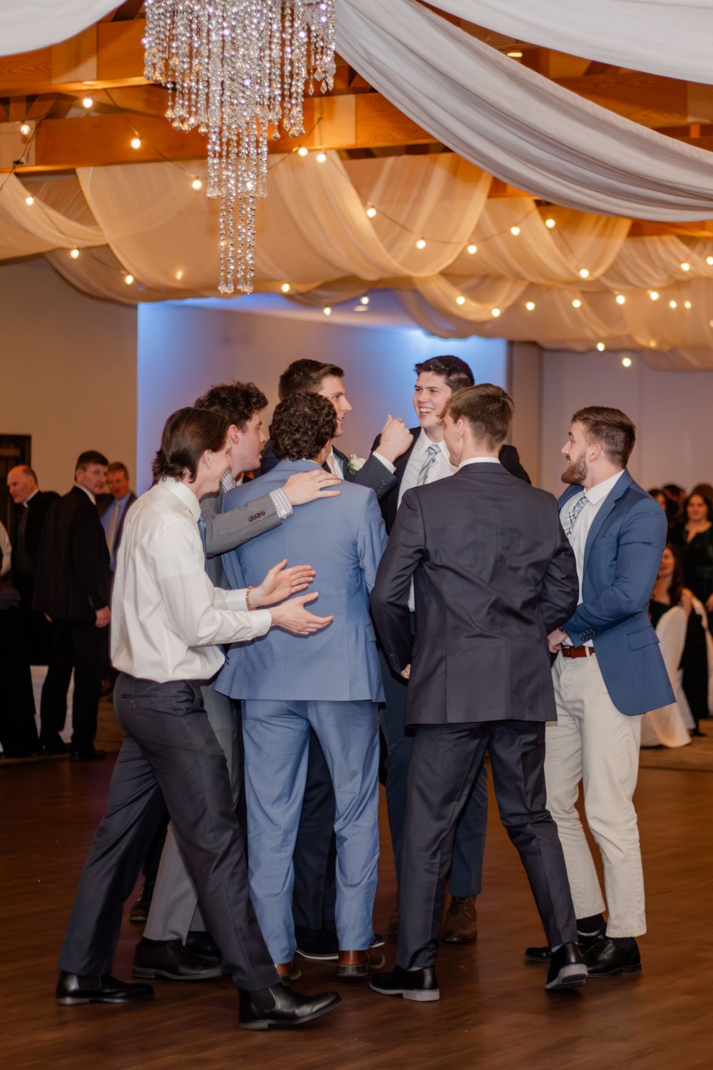Groomsmen in suits huddle together on a decorated wedding reception dance floor with string lights and draped fabric.