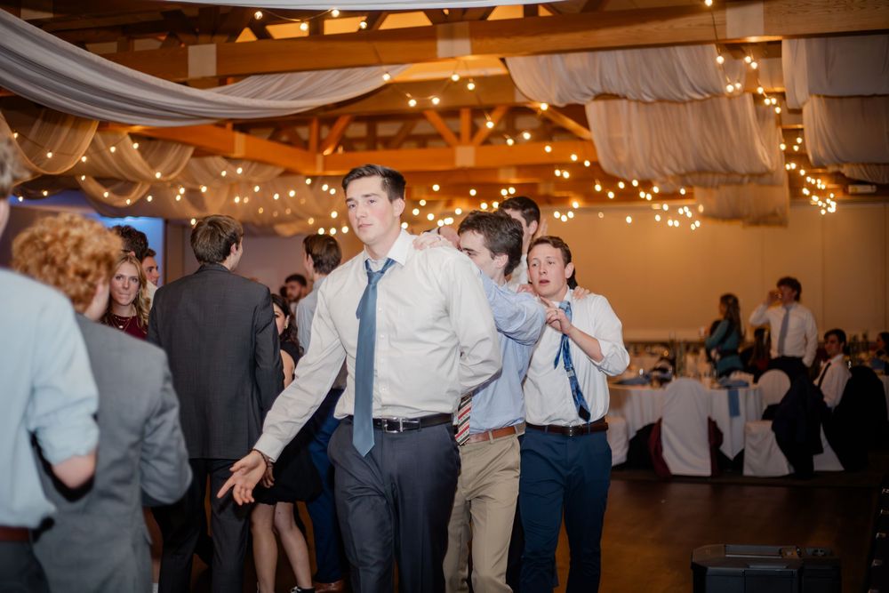 Guests dancing at a wedding reception in a rustic venue decorated with string lights and draped fabric.