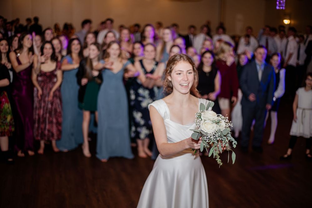 Bride smiles while preparing to toss wedding bouquet to excited guests on dance floor.