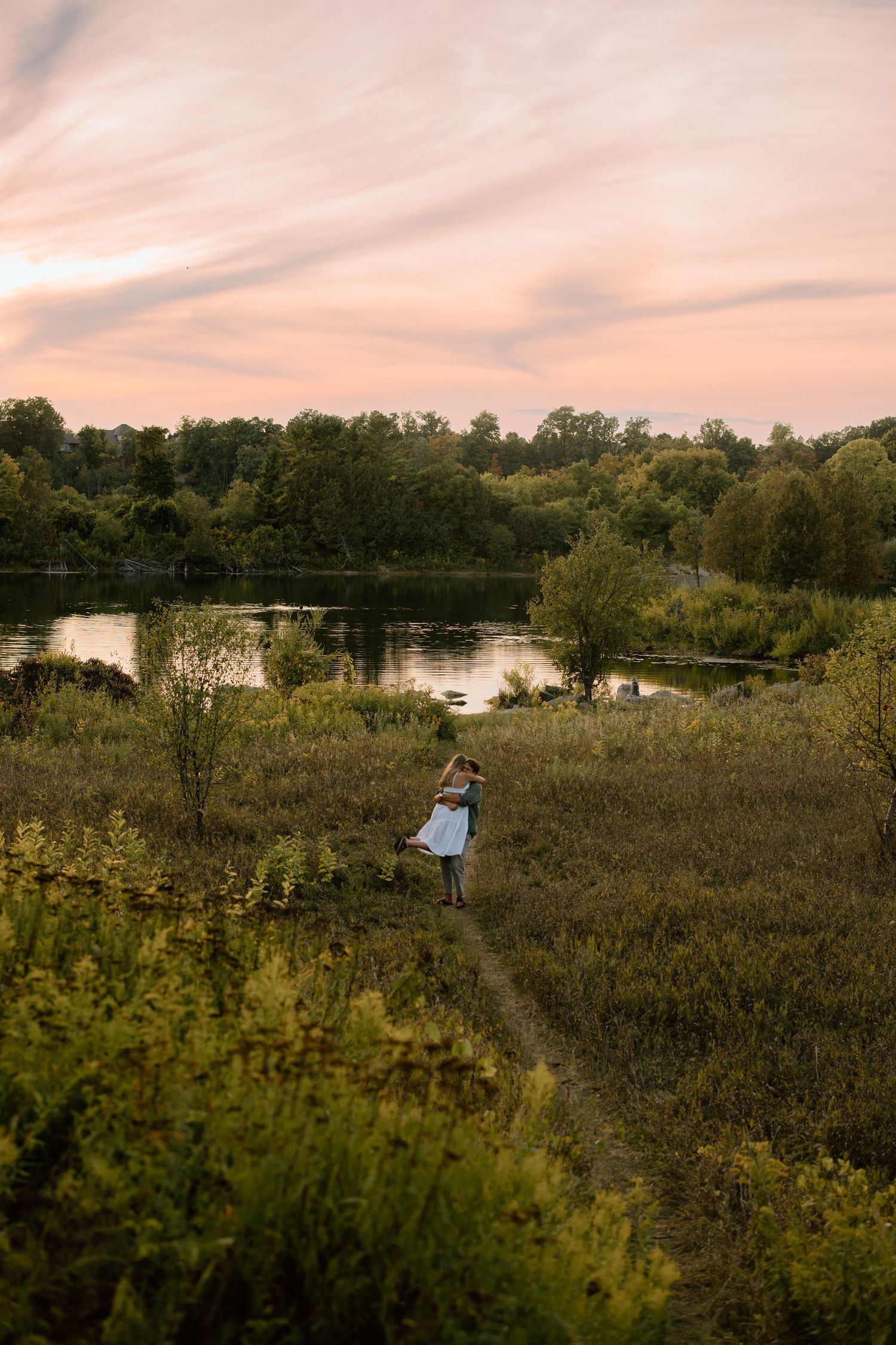 Person walking through grassy field toward lake at sunset with trees and pink sky in background.