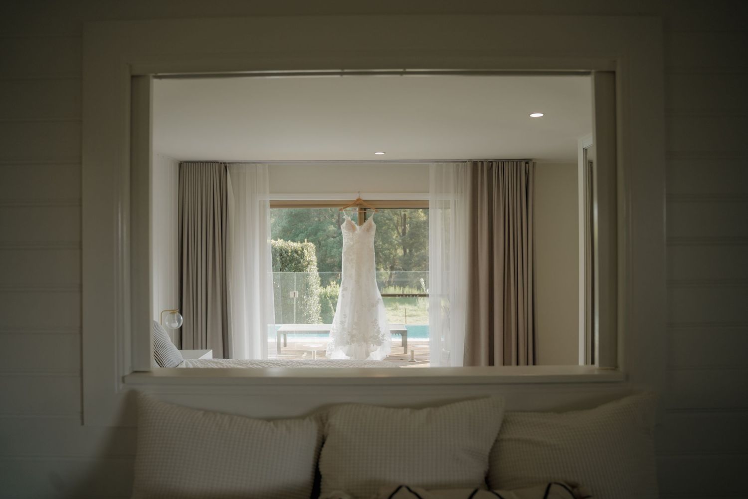 Elegant white wedding dress hanging in a sunlit window with neutral curtains in a bright bridal suite.