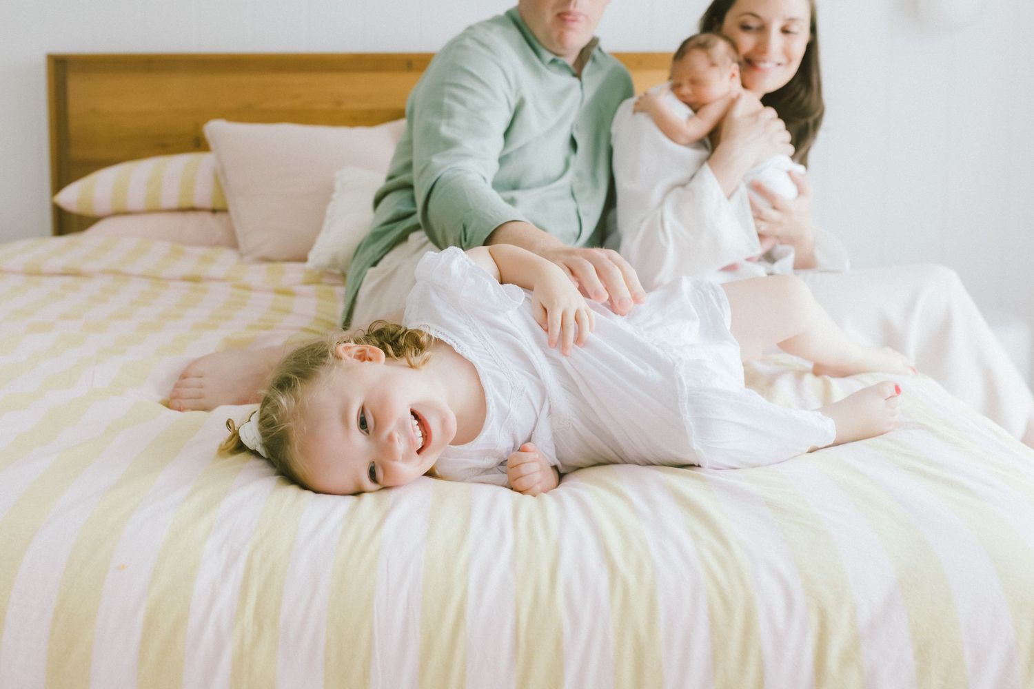 Happy toddler lying on bed laughing while parents hold newborn baby in bright, cozy bedroom setting.