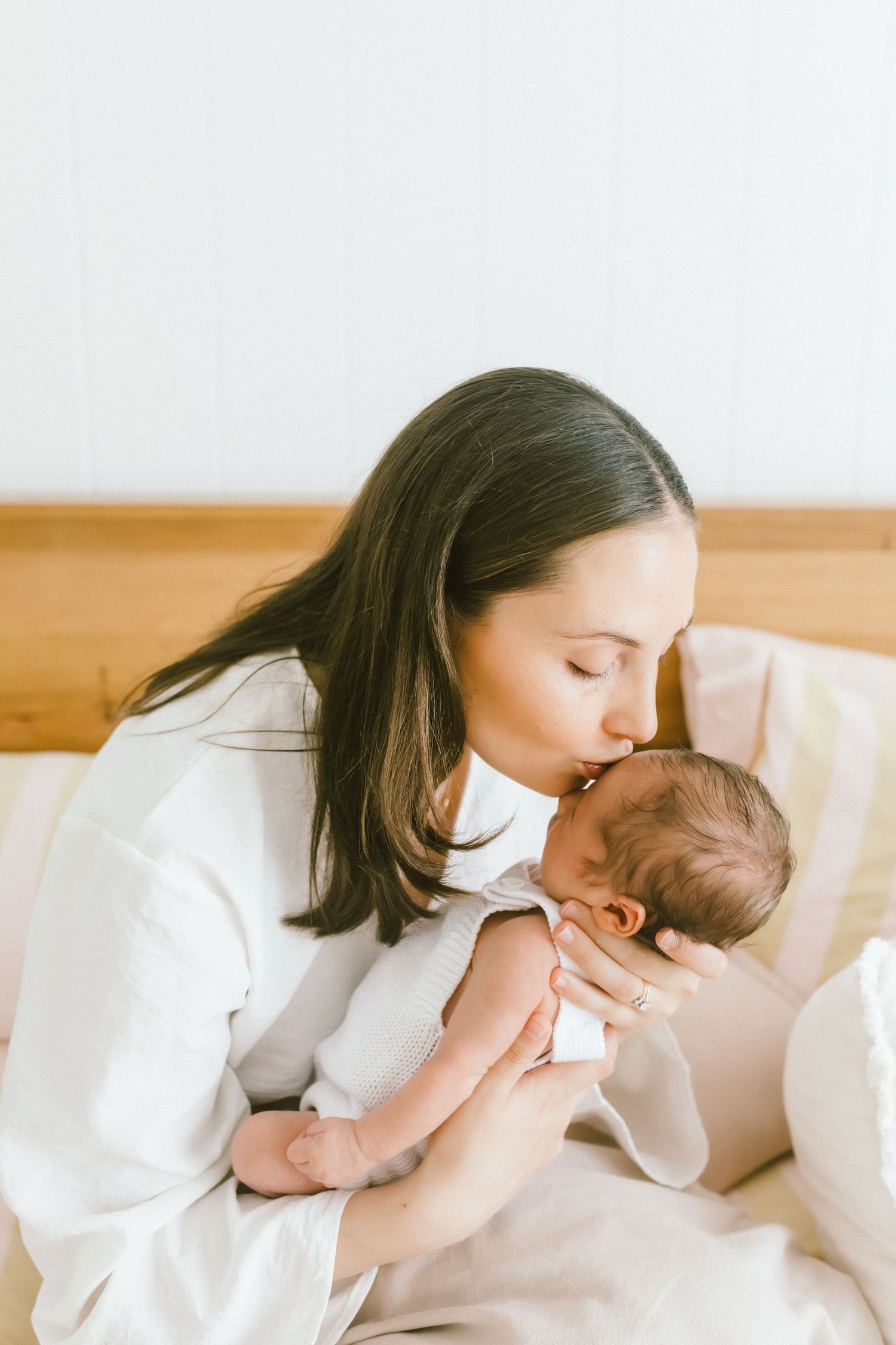 A tender moment as a dark-haired mother in white gently kisses her newborn baby while sitting on a bed.
