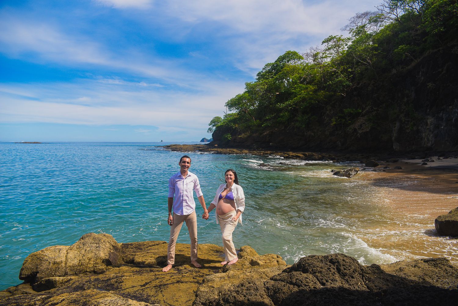 Couple holding hands on rocky beach with turquoise water, lush green cliffs, and clear blue sky in tropical setting.