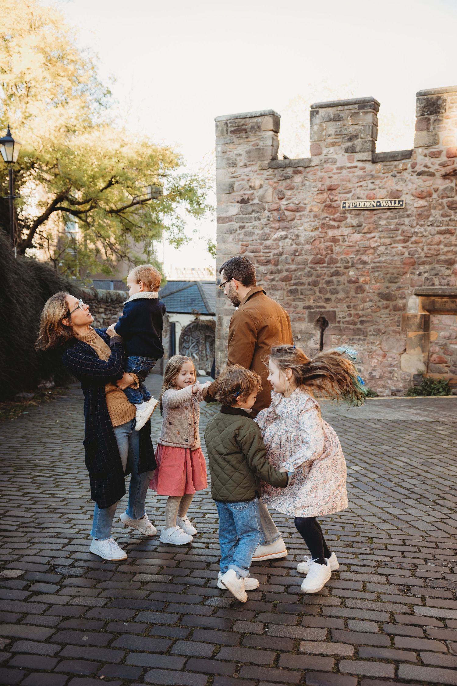 Family with young children playing together on cobblestone street near historic brick wall in autumn.