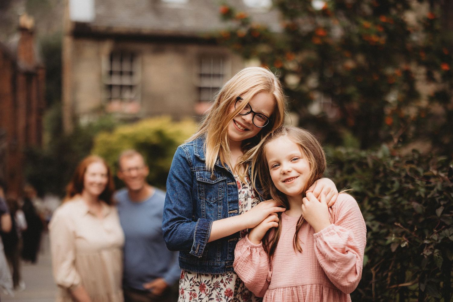 Two smiling girls pose outdoors, older girl with glasses hugging younger girl, family blurred in background.
