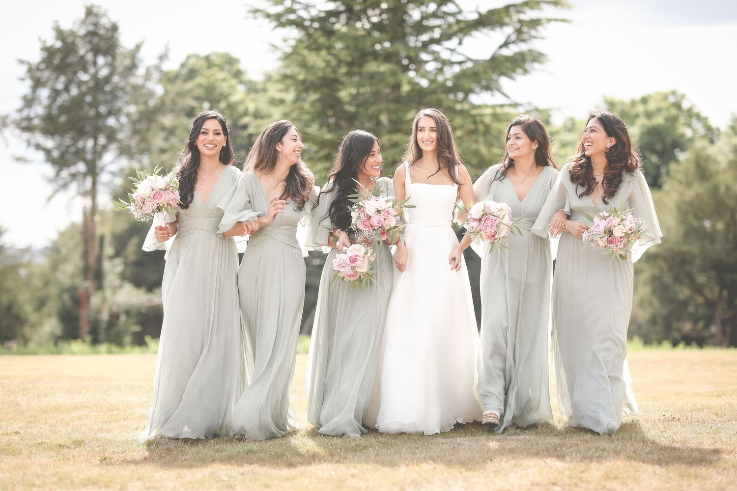 Bride in white gown with five bridesmaids in sage green dresses holding pink bouquets outdoors.