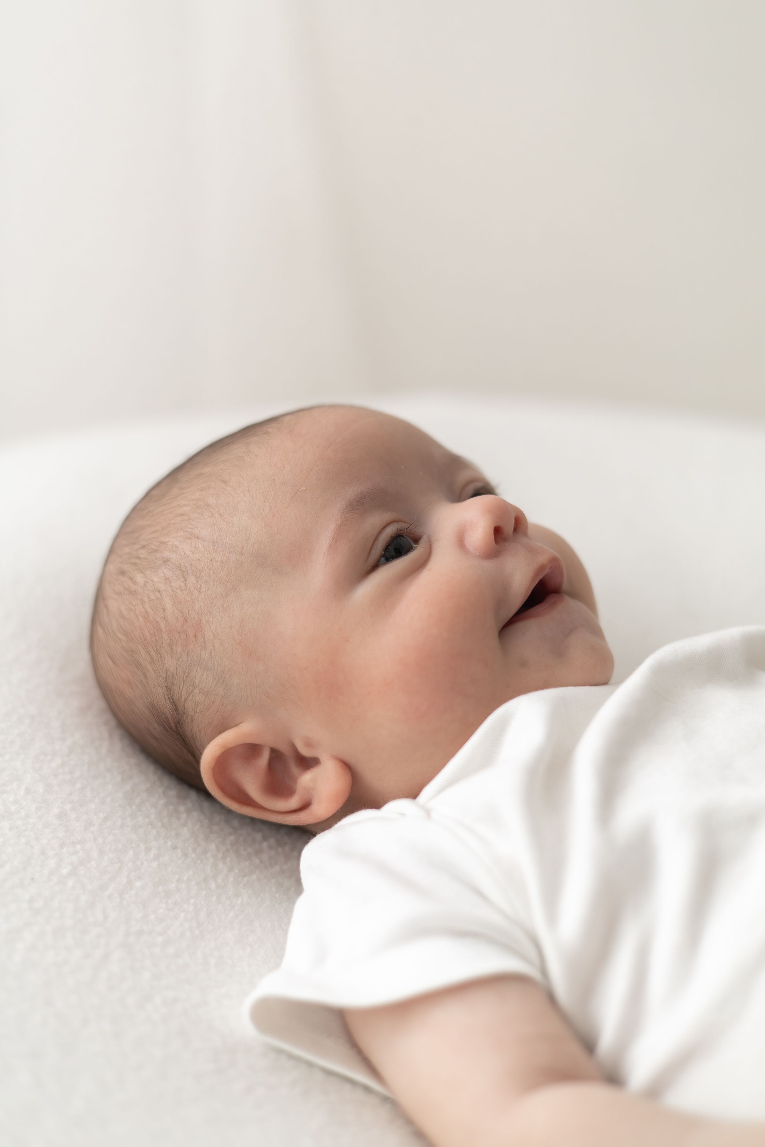 Smiling newborn baby lying on a white surface wearing a white onesie, looking upward older newborn photographer Nottingham cosy home studio
