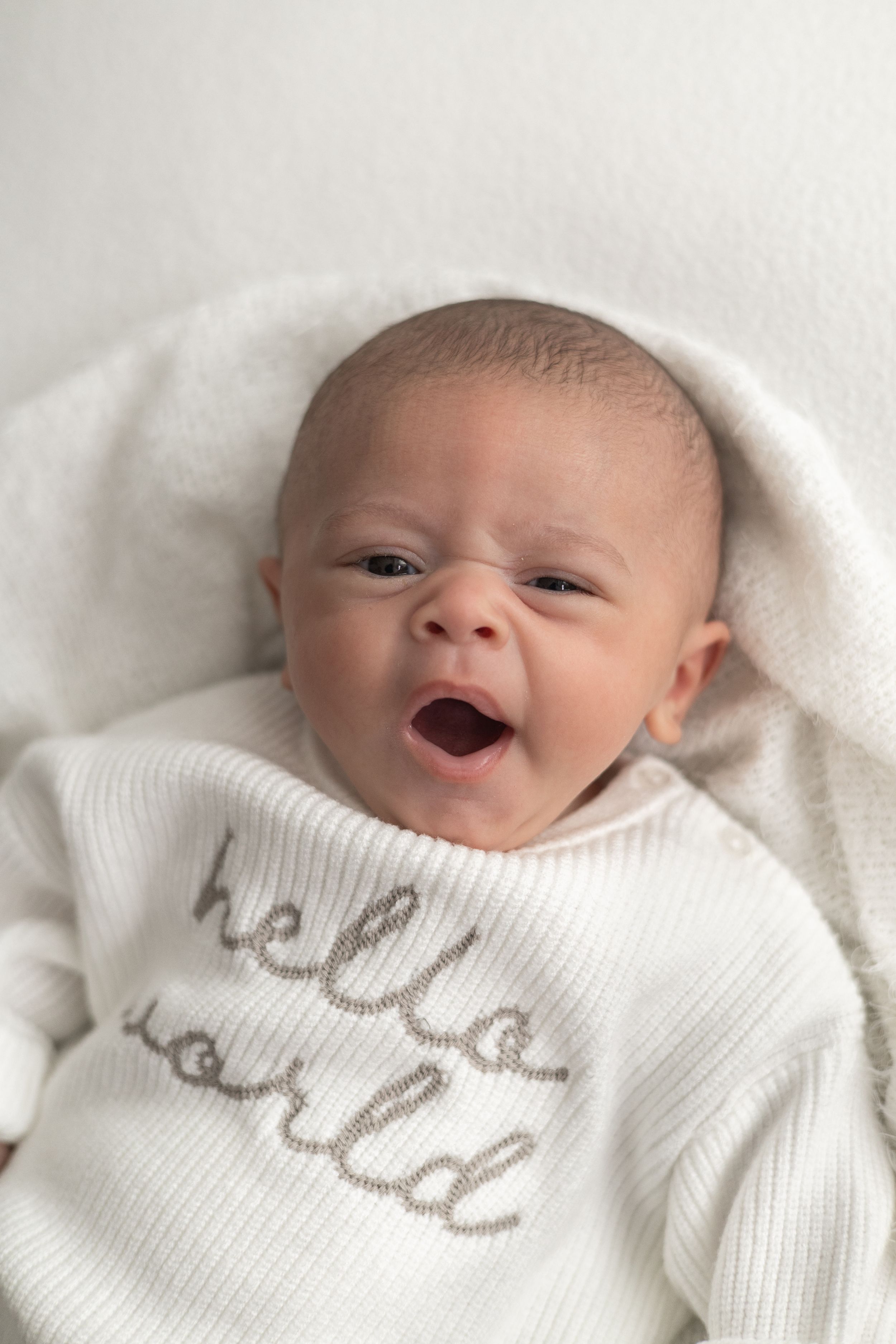Newborn baby yawning in a white 'hello world' onesie, wrapped in a soft white blanket older newborn photographer Nottingham cosy home studio