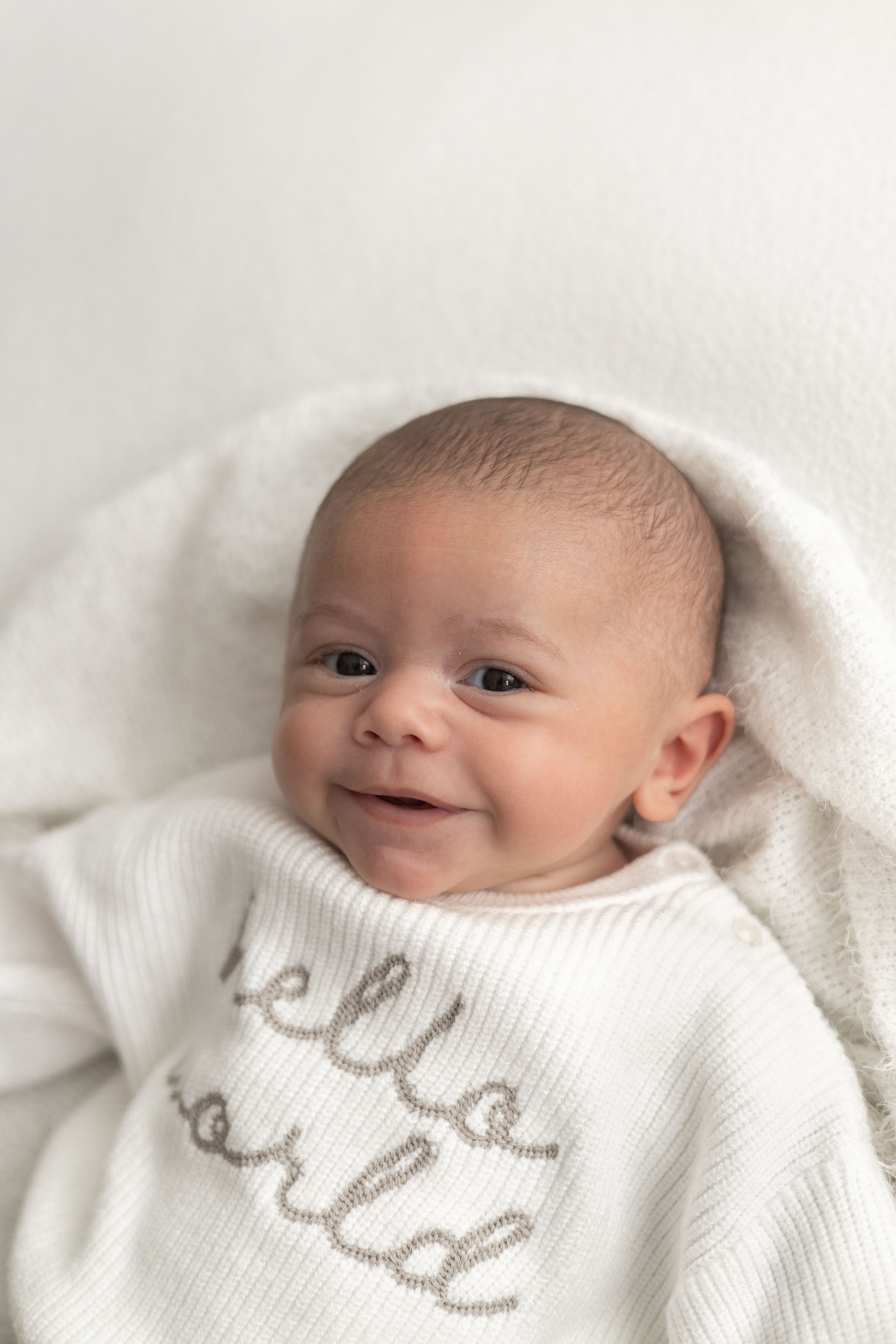 Smiling newborn baby wearing in a white “Hello World” romper, lying on a soft white background older newborn photographer Nottingham studio