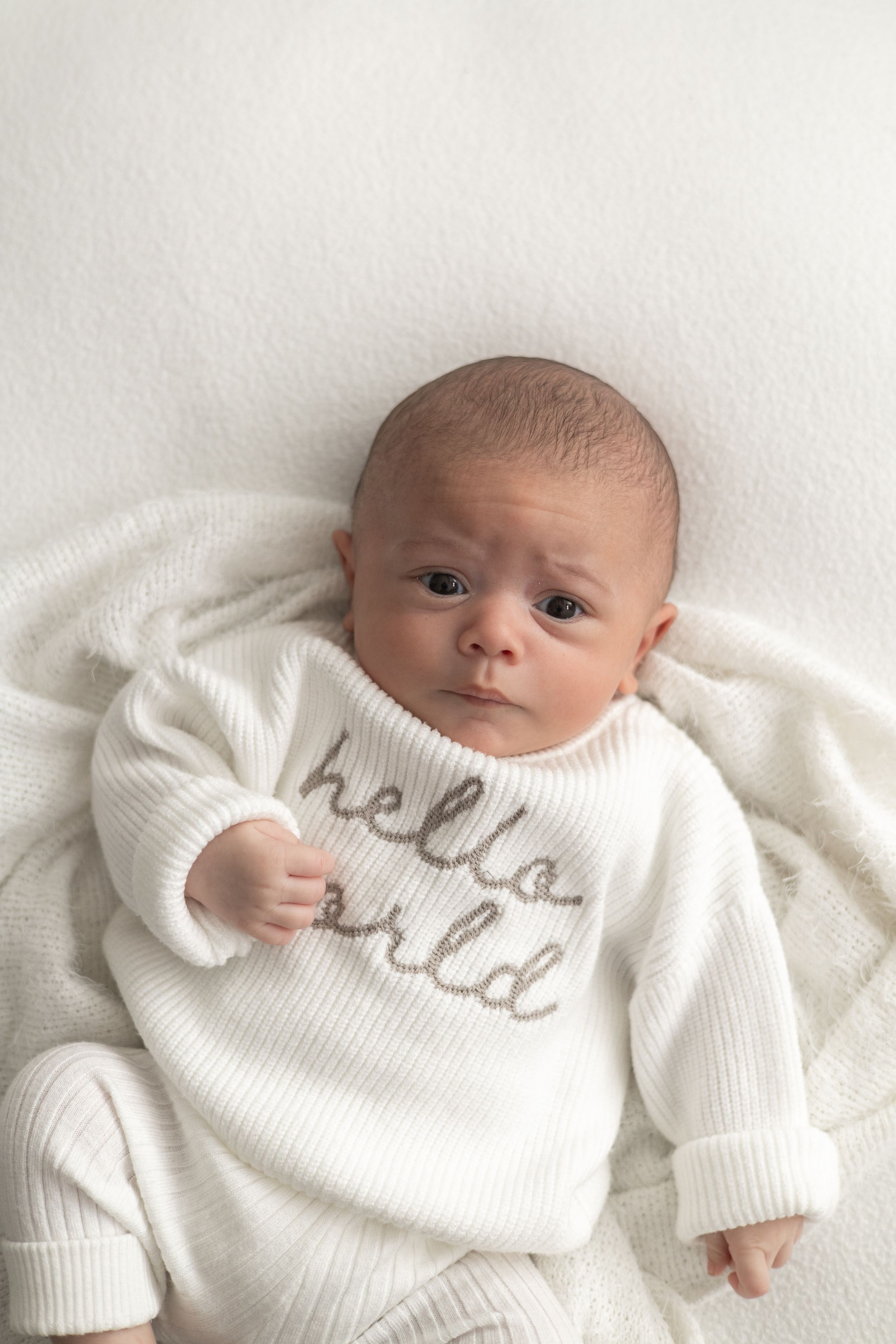 Newborn baby lying on white blanket wearing a cream knit 'hello world' romper older newborn photographer Nottingham cosy home studio