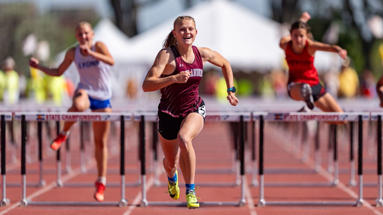 Female athlete smiling while clearing a hurdle during a track and field championship race, competitors visible behind her.
