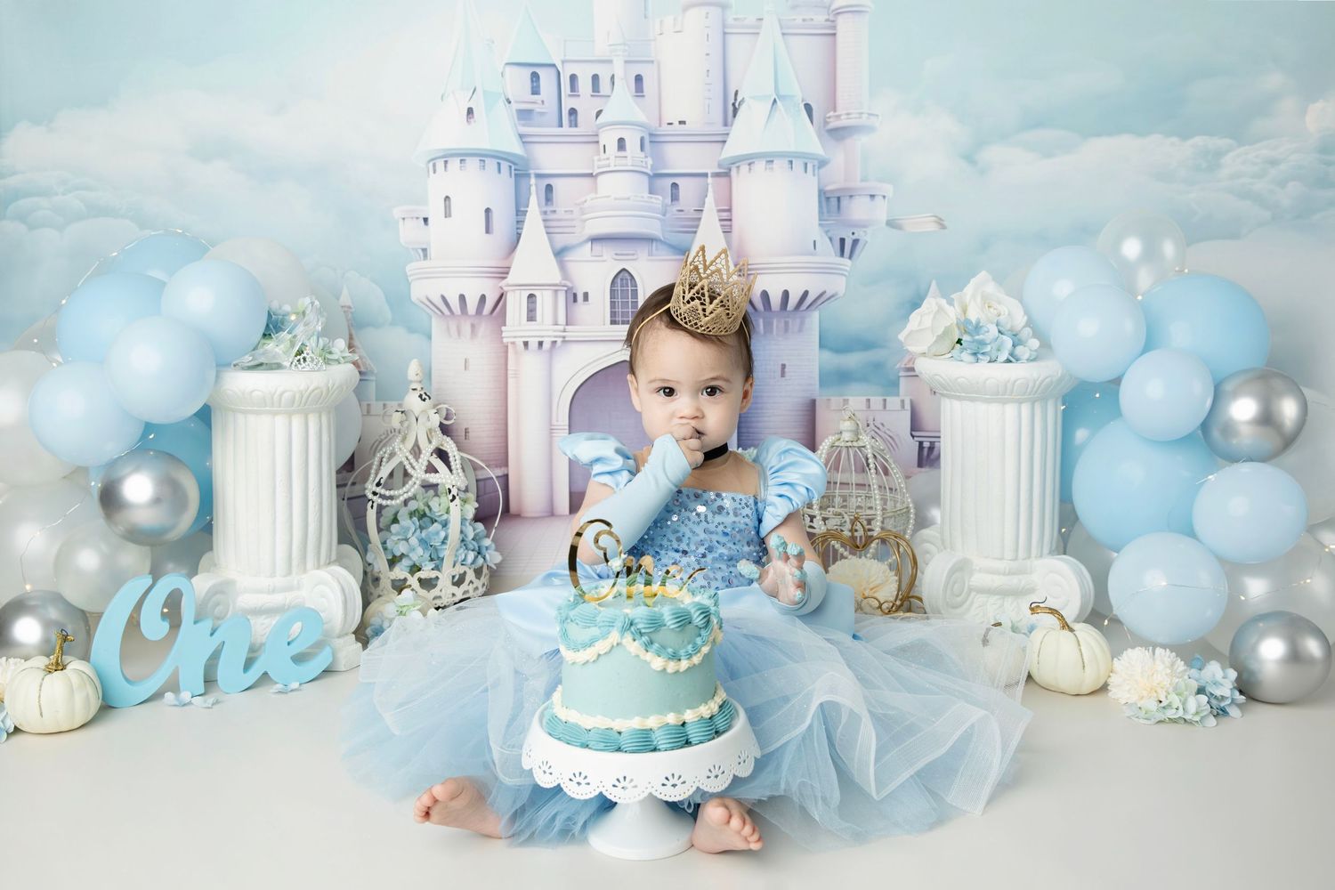 Baby in blue dress sits before castle backdrop with balloons, wearing crown, touching first birthday cake.