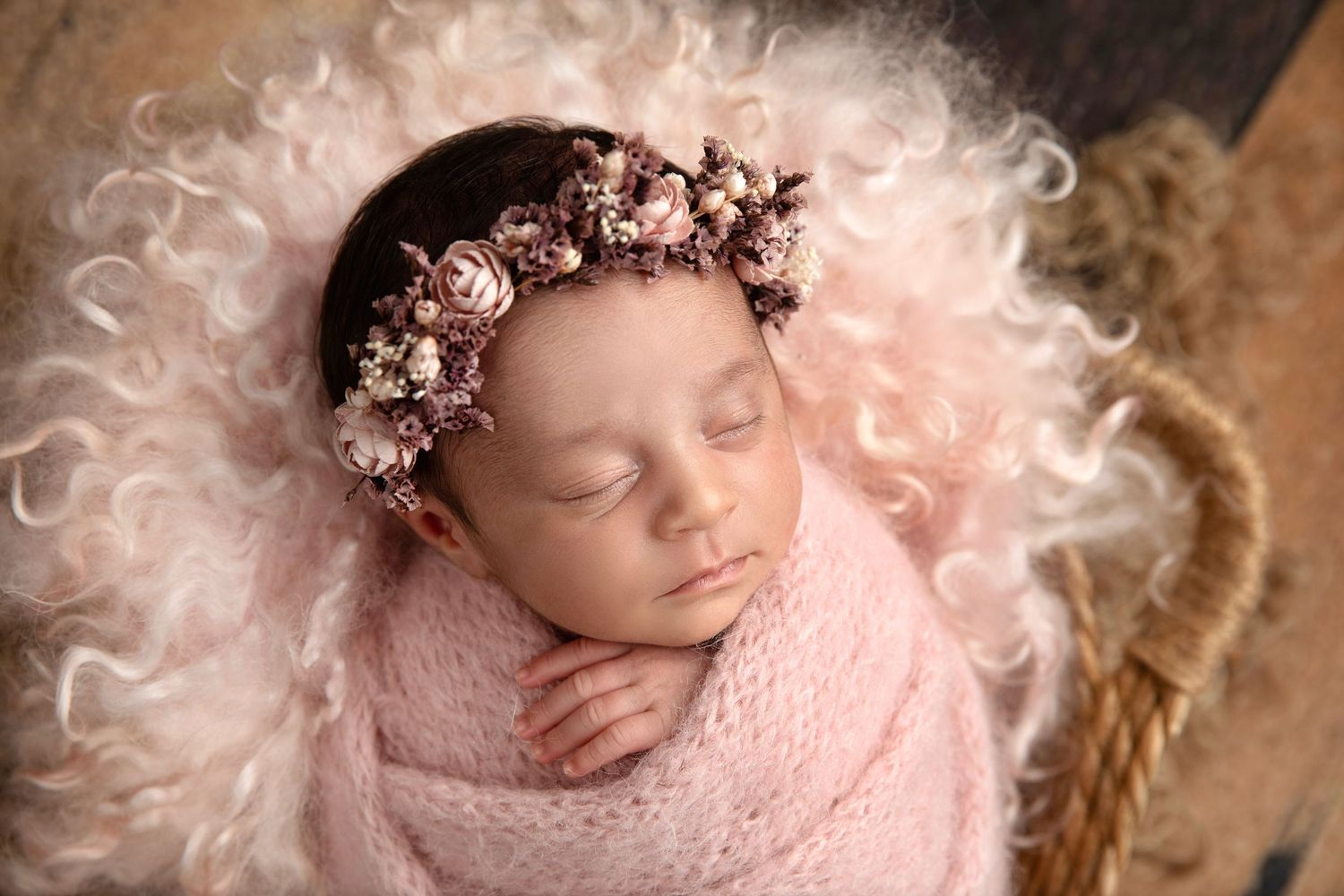 Sleeping newborn baby wrapped in pink with a floral crown, nestled in a fluffy basket for a professional photo.