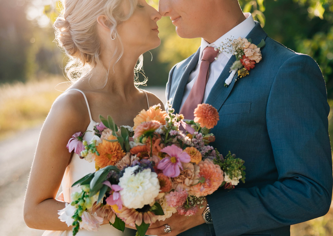 A bride and groom share a romantic moment holding a vibrant bouquet of pink, orange, and white flowers outdoors.