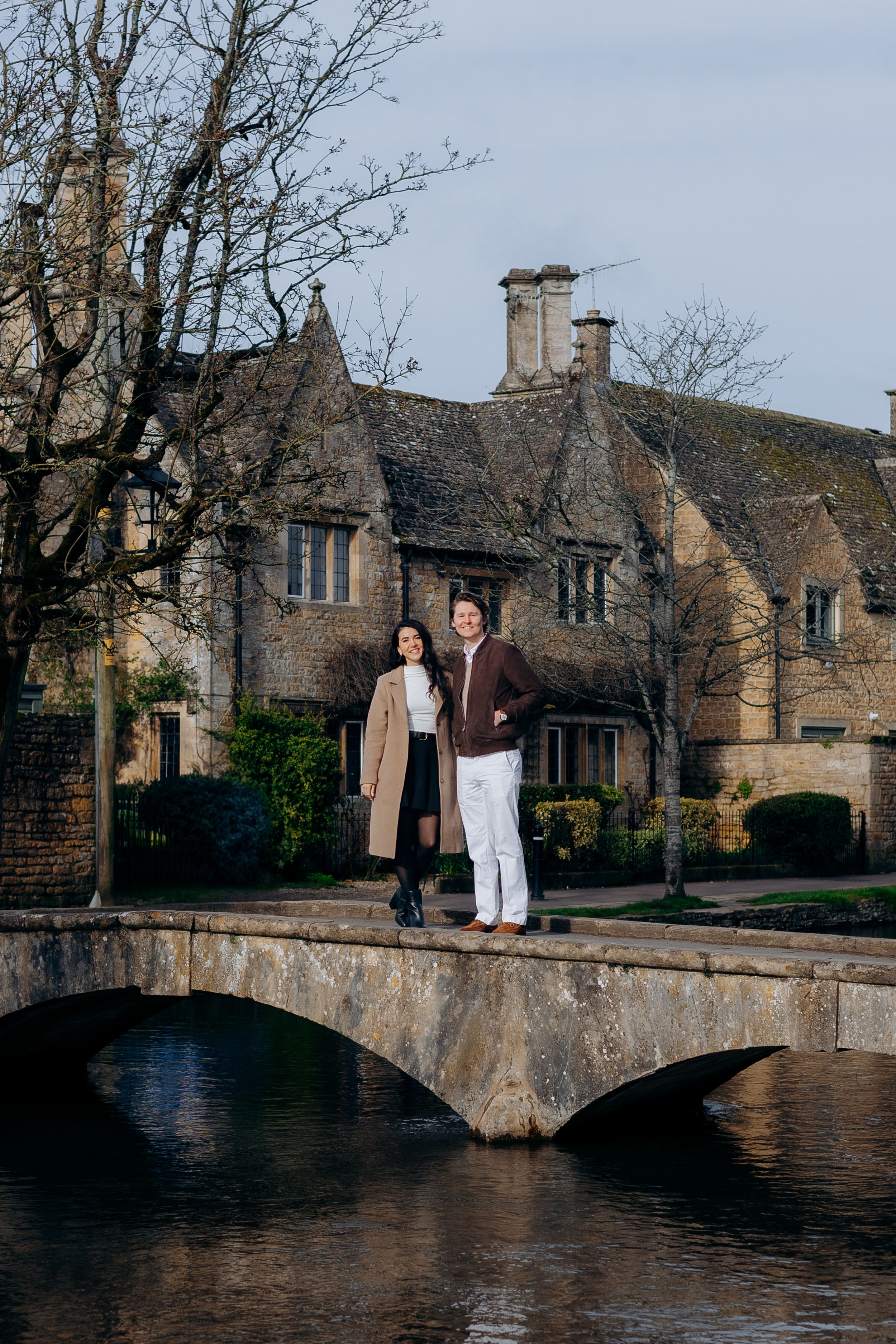 Couple standing on a stone bridge over a river in the Cotswolds, England, with historic honey-colored stone cottages behind them at Bourton.