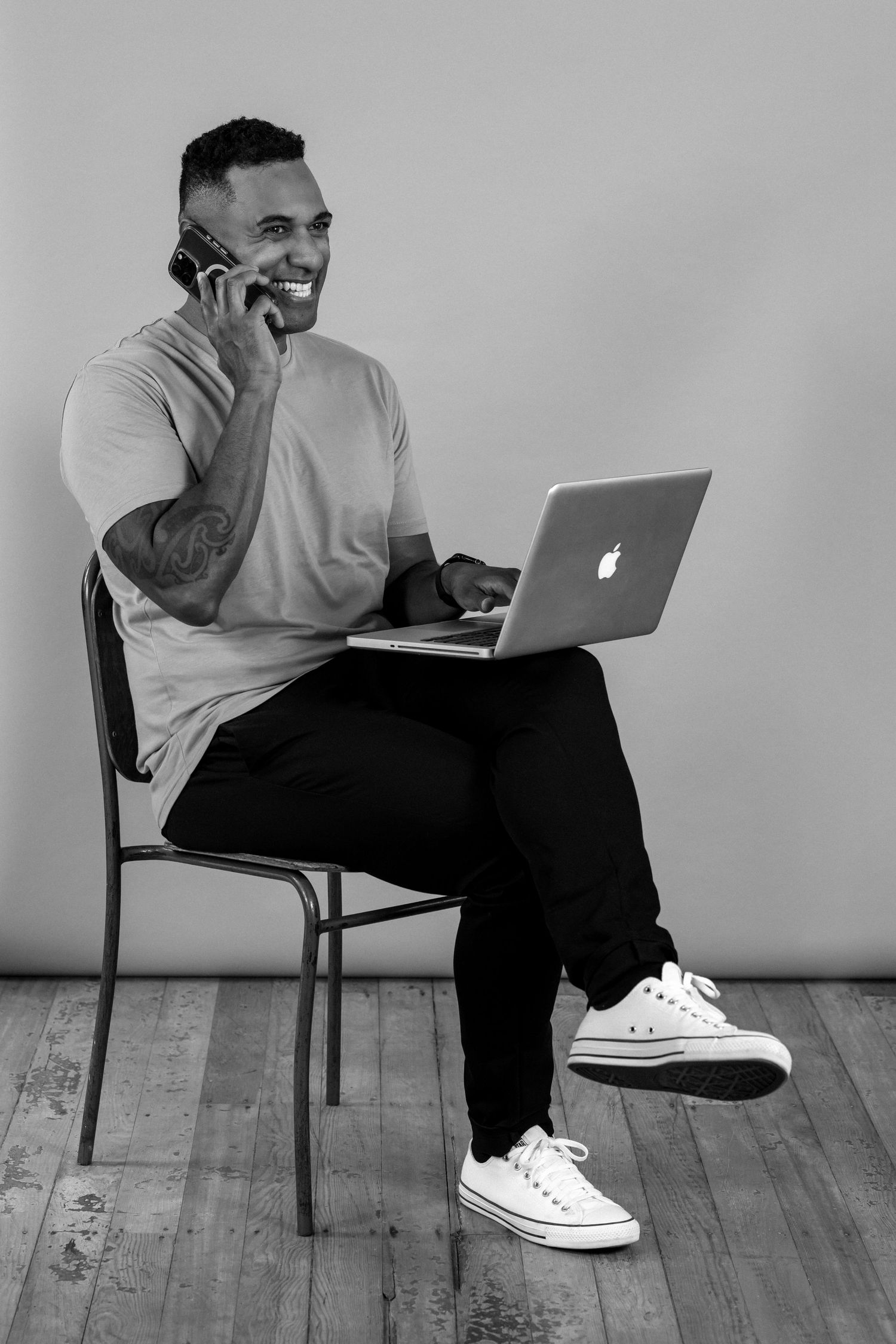 Black and white image of a man sitting down crossed legged with a laptop on his lap. He is talking on the phone and smiling. Branding