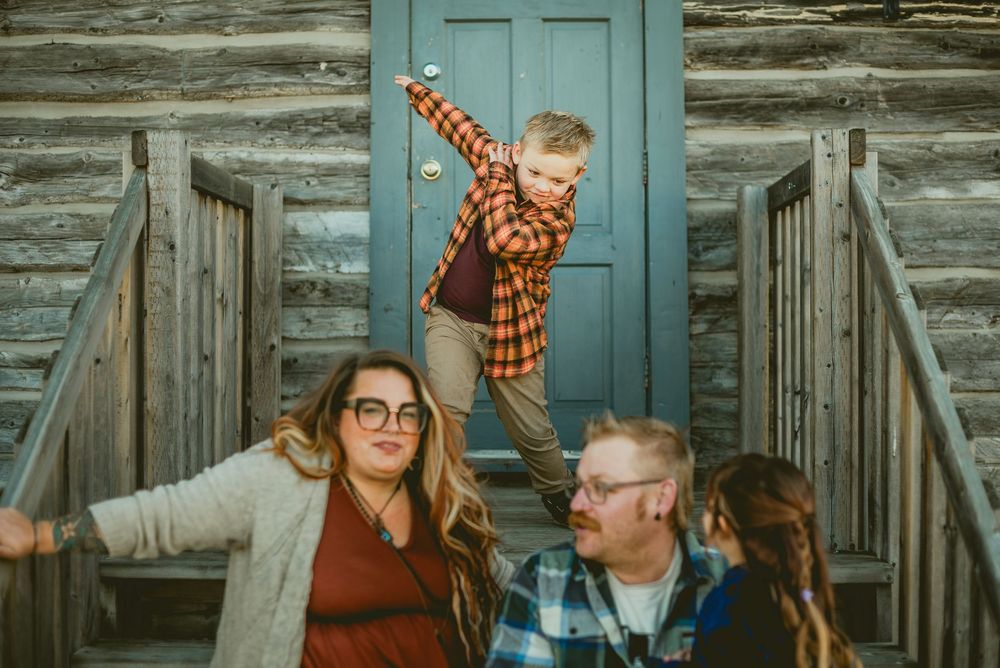 Family of four posing on cabin steps, boy balancing on railing, parents smiling, rustic log wall backdrop.