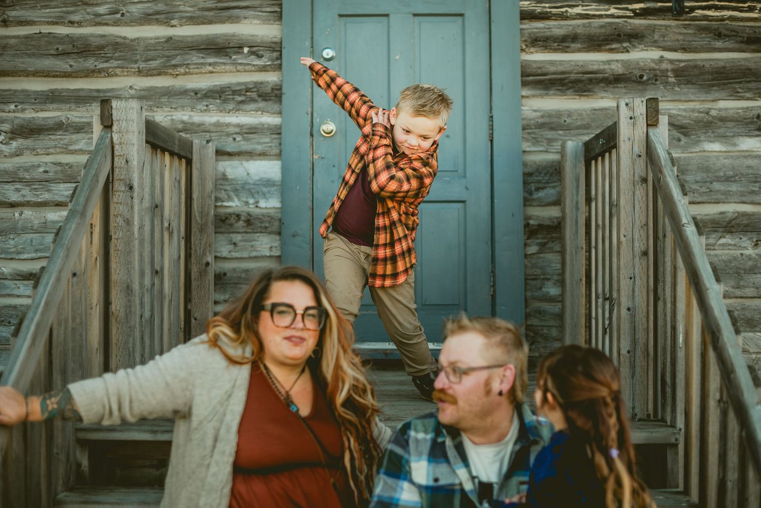 Family of four posing on cabin steps, boy balancing on railing, parents smiling, rustic log wall backdrop.