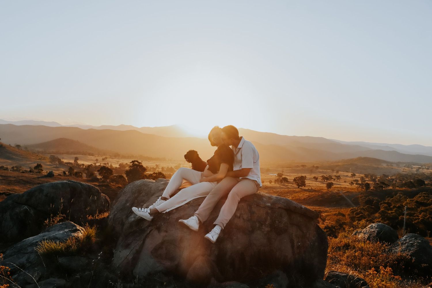 Couple sitting on rocks sharing a romantic kiss during golden sunset with scenic valley landscape in background.