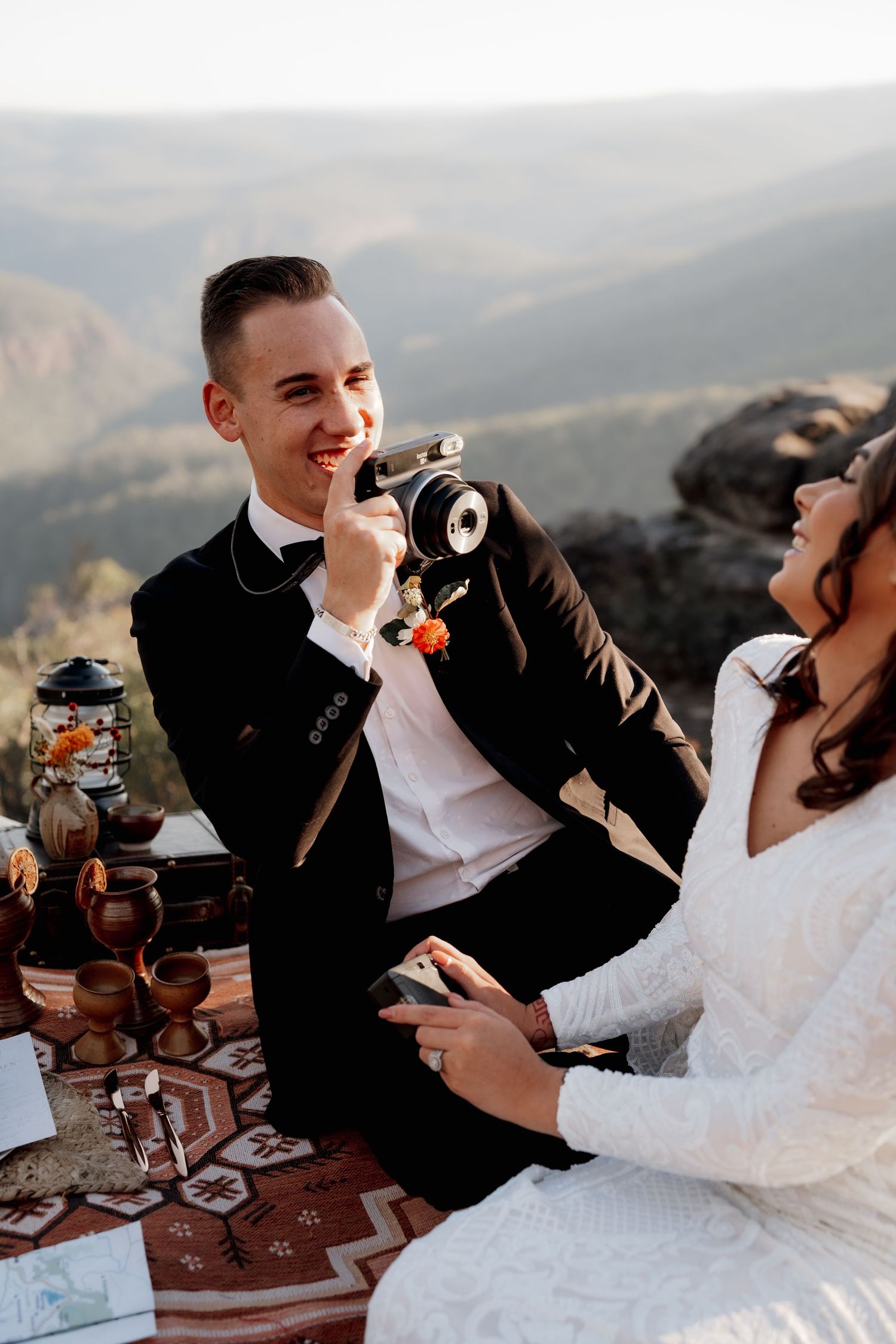 Groom in black tuxedo holds vintage camera while bride in white lace dress sits together at mountain elopement picnic.