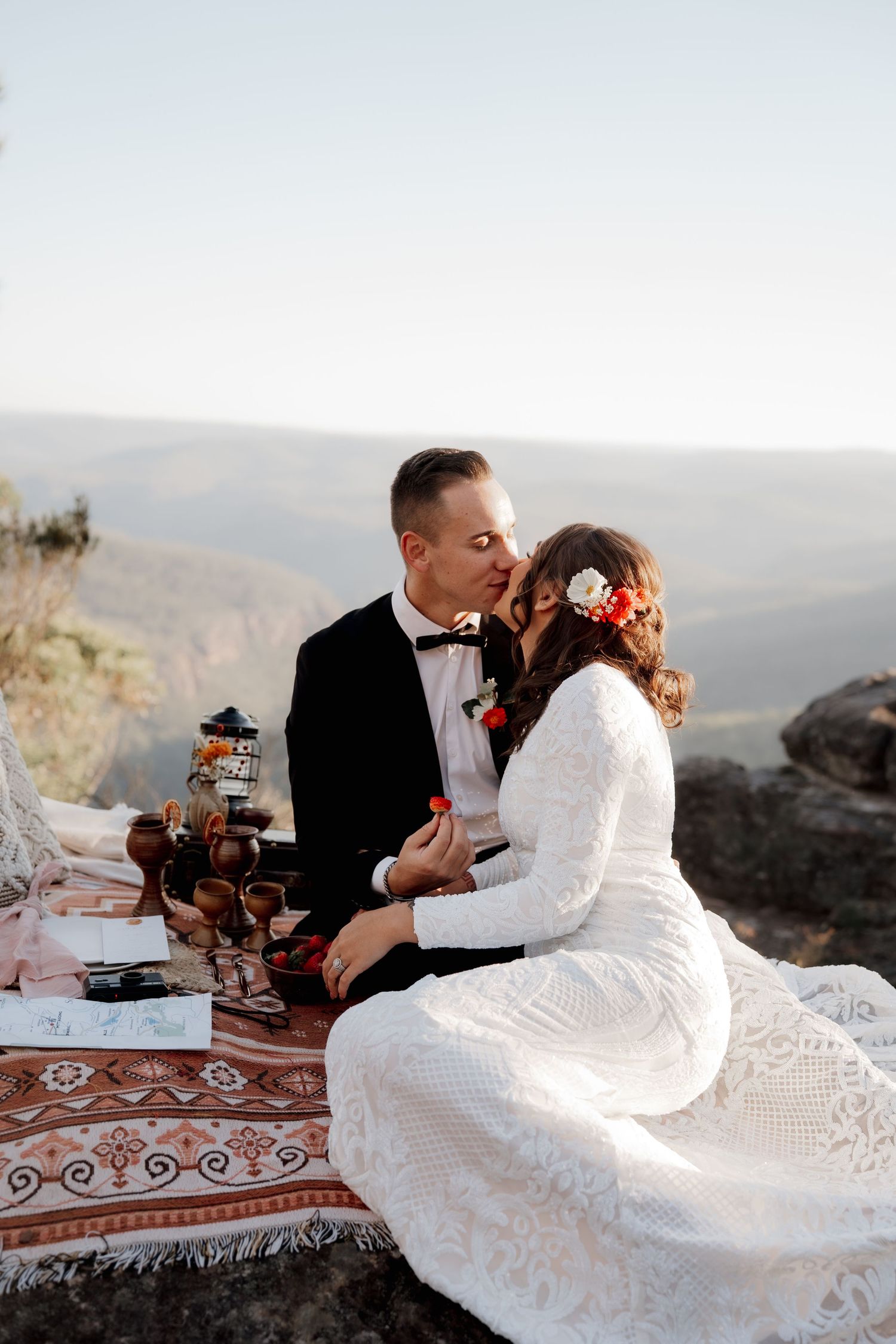 Bride and groom share a kiss during an outdoor mountain elopement picnic, bride wearing lace dress with floral hair accessories.