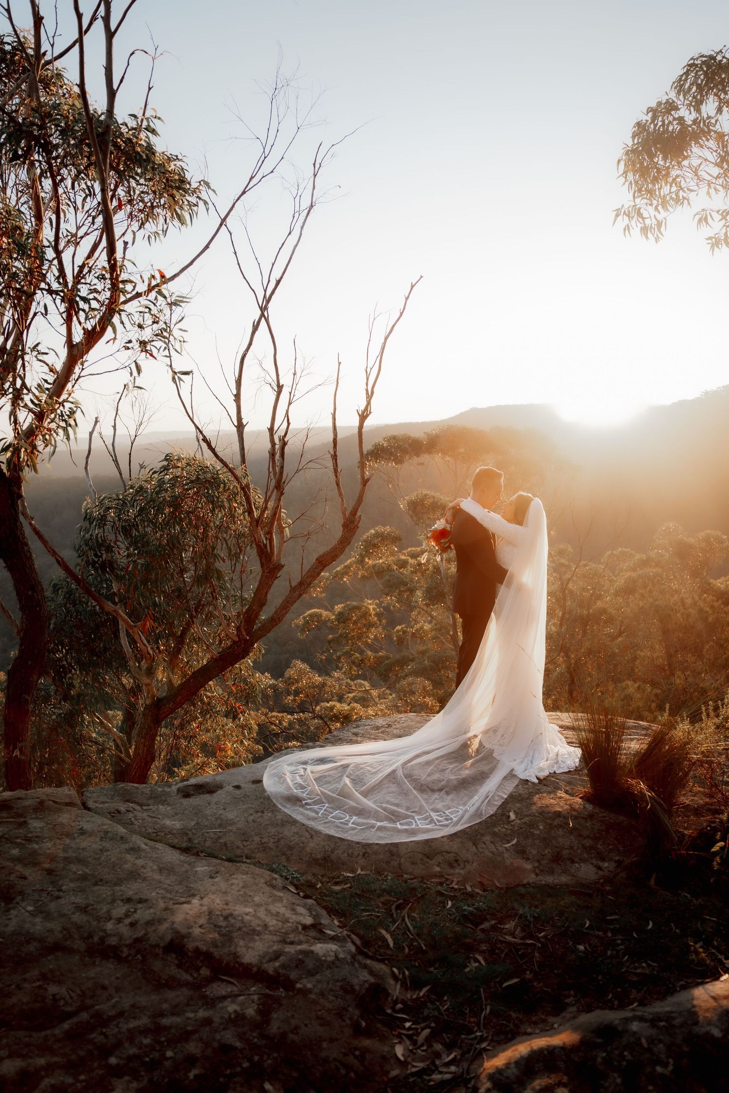 Bride and groom embrace on rocky cliff at golden sunset with flowing lace veil and mountain views in Australia.