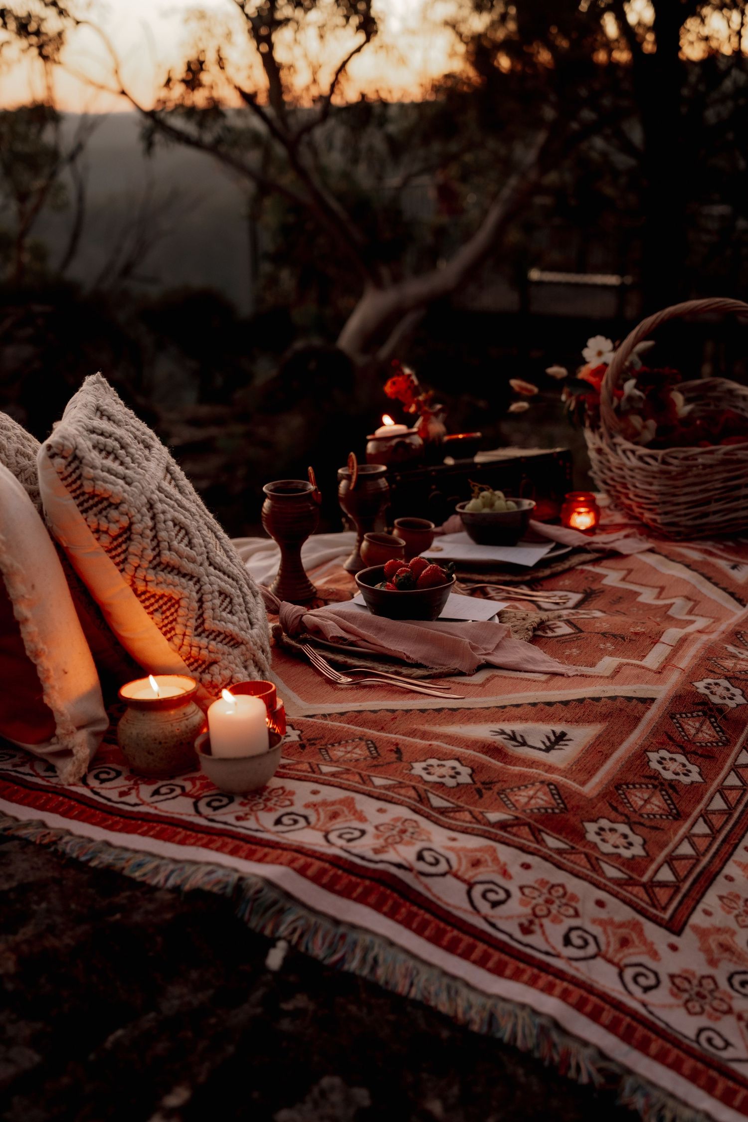 Romantic boho outdoor picnic setup at dusk with candles, patterned blanket, pillows, and fruit on a rustic table.